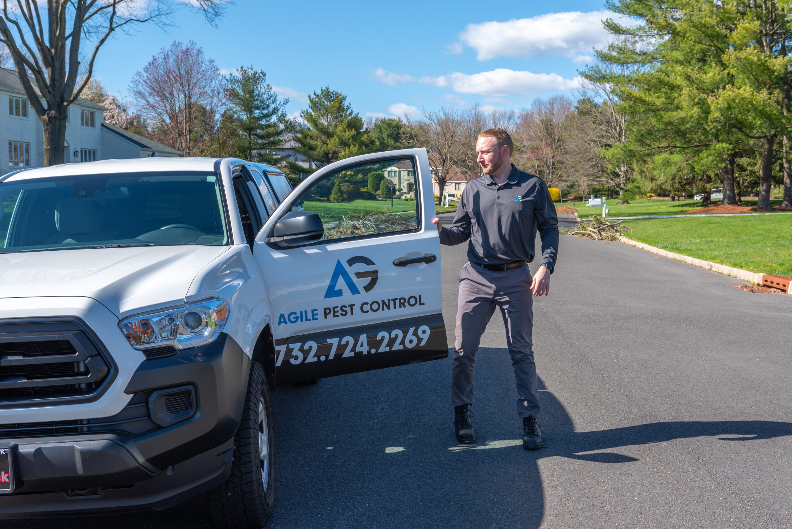 Agile Pest Control technician in uniform servicing a property in Colts Neck, NJ, with a branded service truck parked nearby.